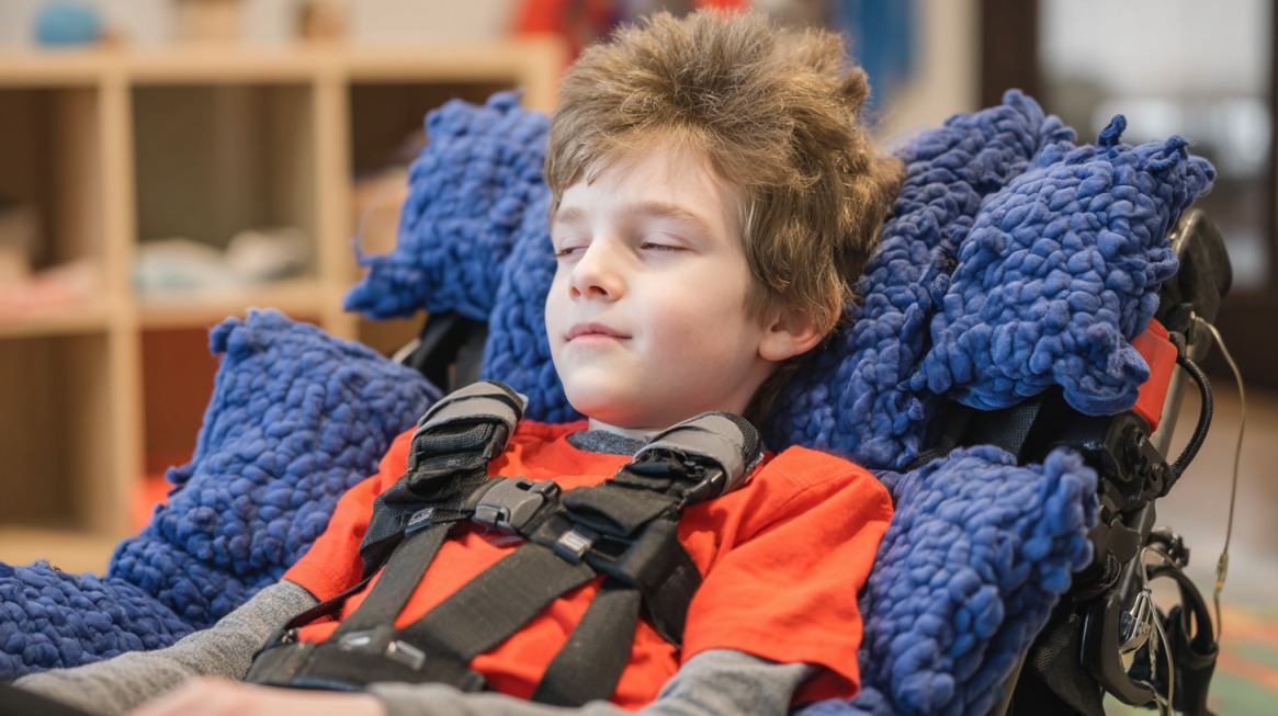 A child resting comfortably in a wheelchair with soft blue cushions and wearing a red shirt, appearing calm and relaxed
