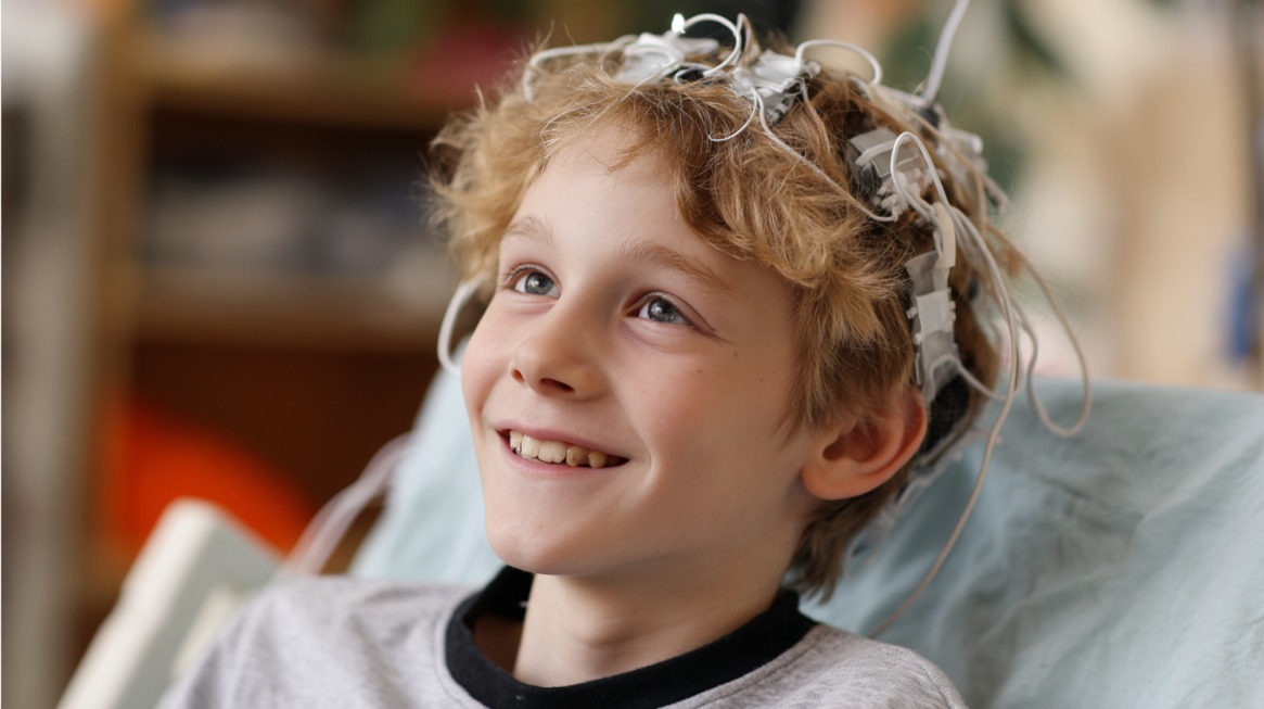 A smiling young child with electrodes attached to their head, likely undergoing a neurological test or EEG, sitting comfortably in a medical setting