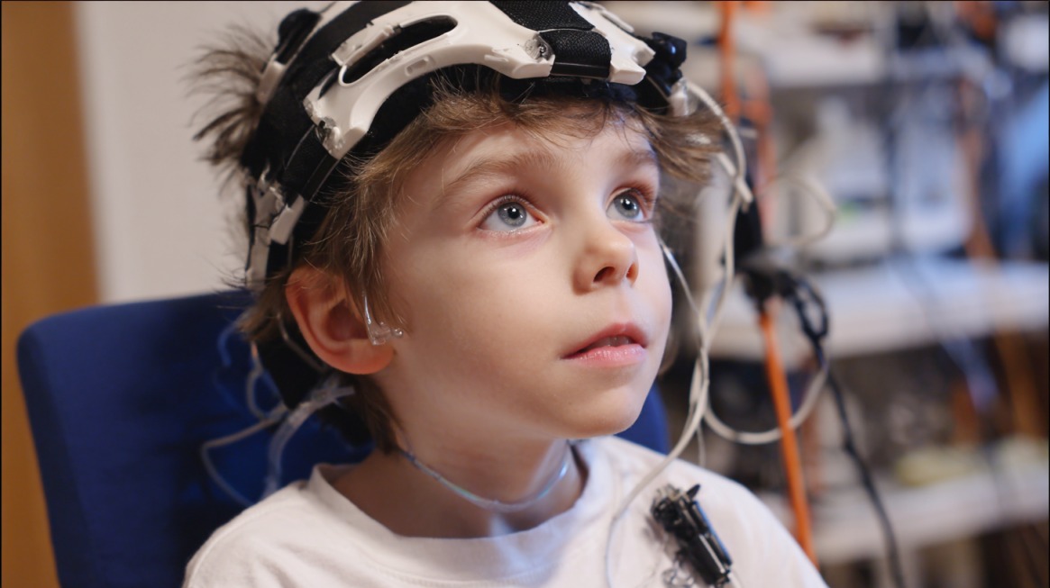 A young child undergoing a neurological study, wearing a white shirt and a head cap with multiple sensors and wires attached, in a medical or research setting