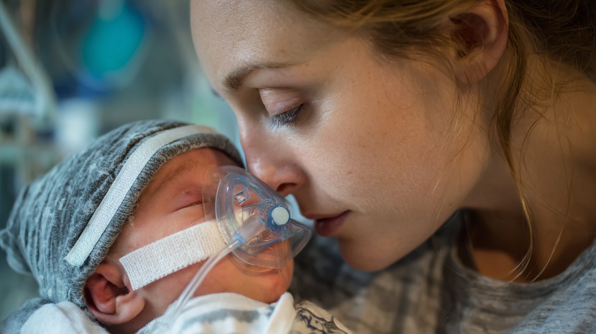 Mother gently touching her nose to her newborn baby’s face; the baby is wearing a hat and connected to an oxygen mask in a hospital setting