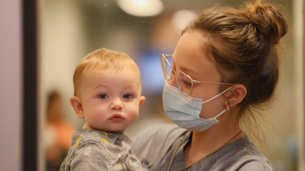 A healthcare professional wearing glasses and a face mask holds a baby in her arms, with the baby looking toward the camera in a softly lit clinic setting
