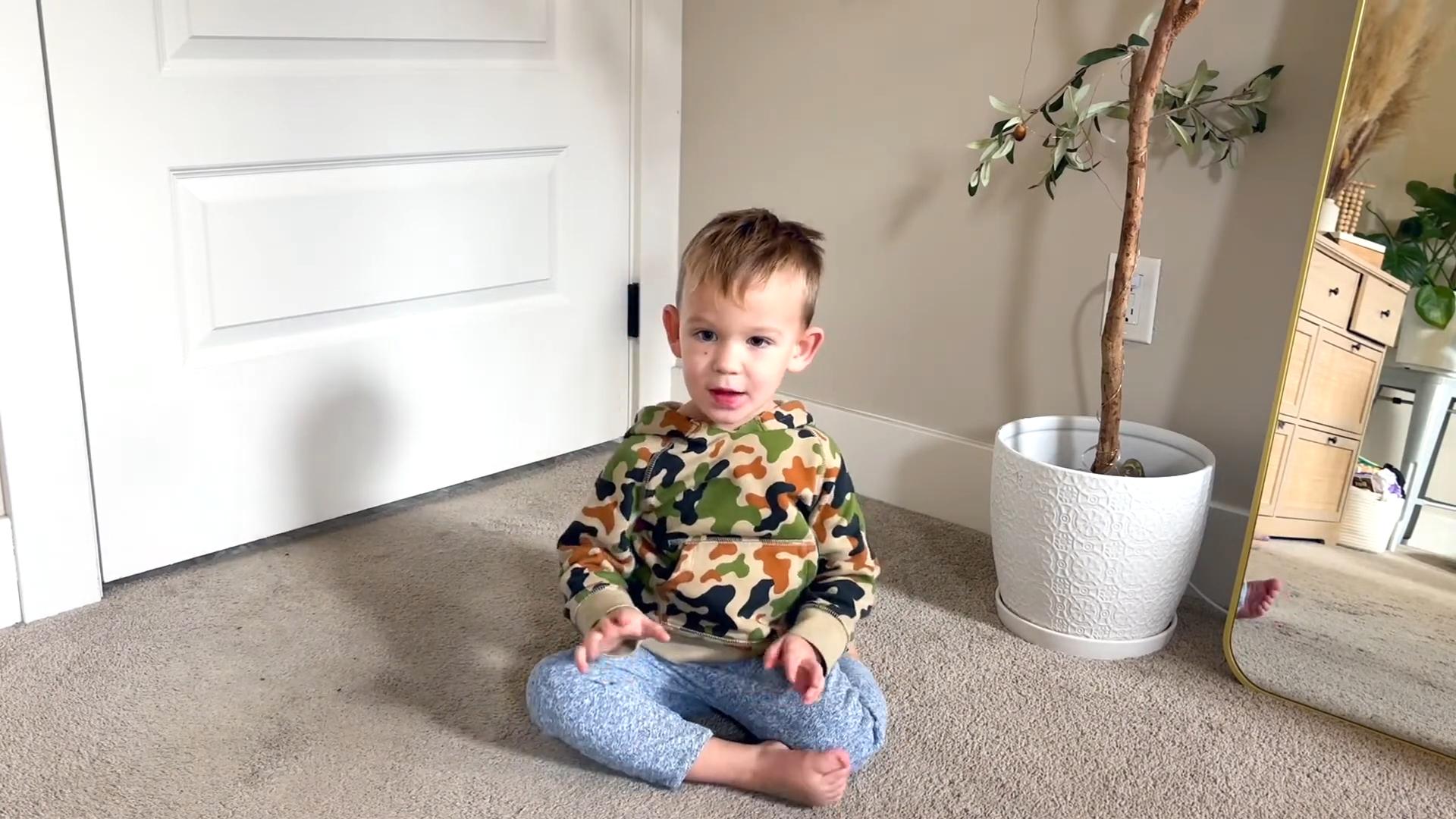 Young child sitting cross legged on carpet indoors near a plant and mirror