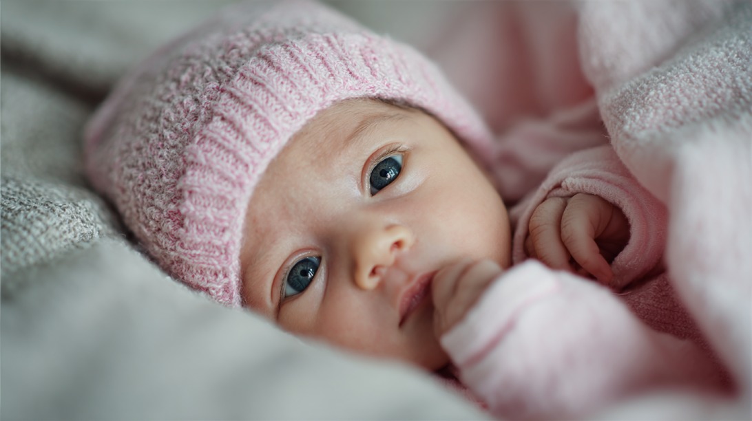 Close up of a baby with blue eyes lying on a blanket and dressed in a pink hat and outfit