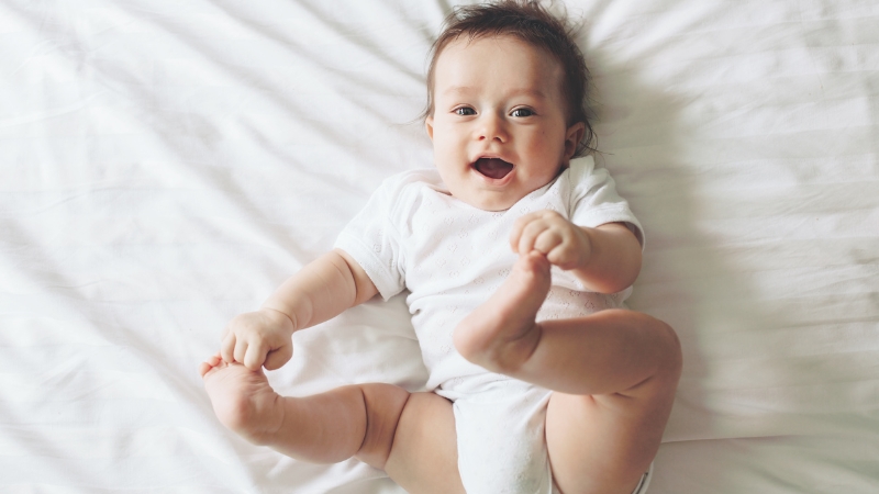 6-month-old baby lying on the bed and holding their feet while smiling