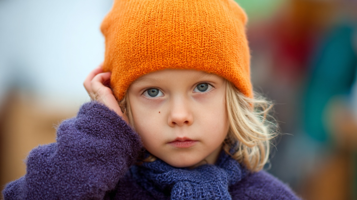 A young child wearing an orange beanie and purple sweater, looking forward while touching their ear