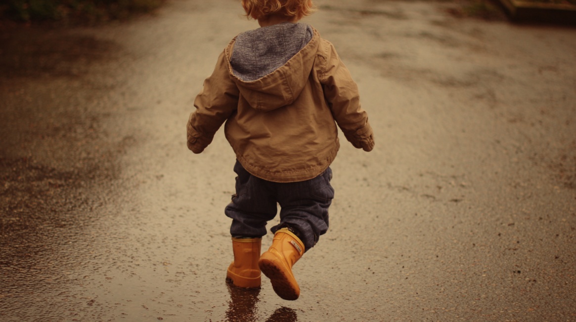 A toddler walking away on a wet path wearing a jacket and rain boots